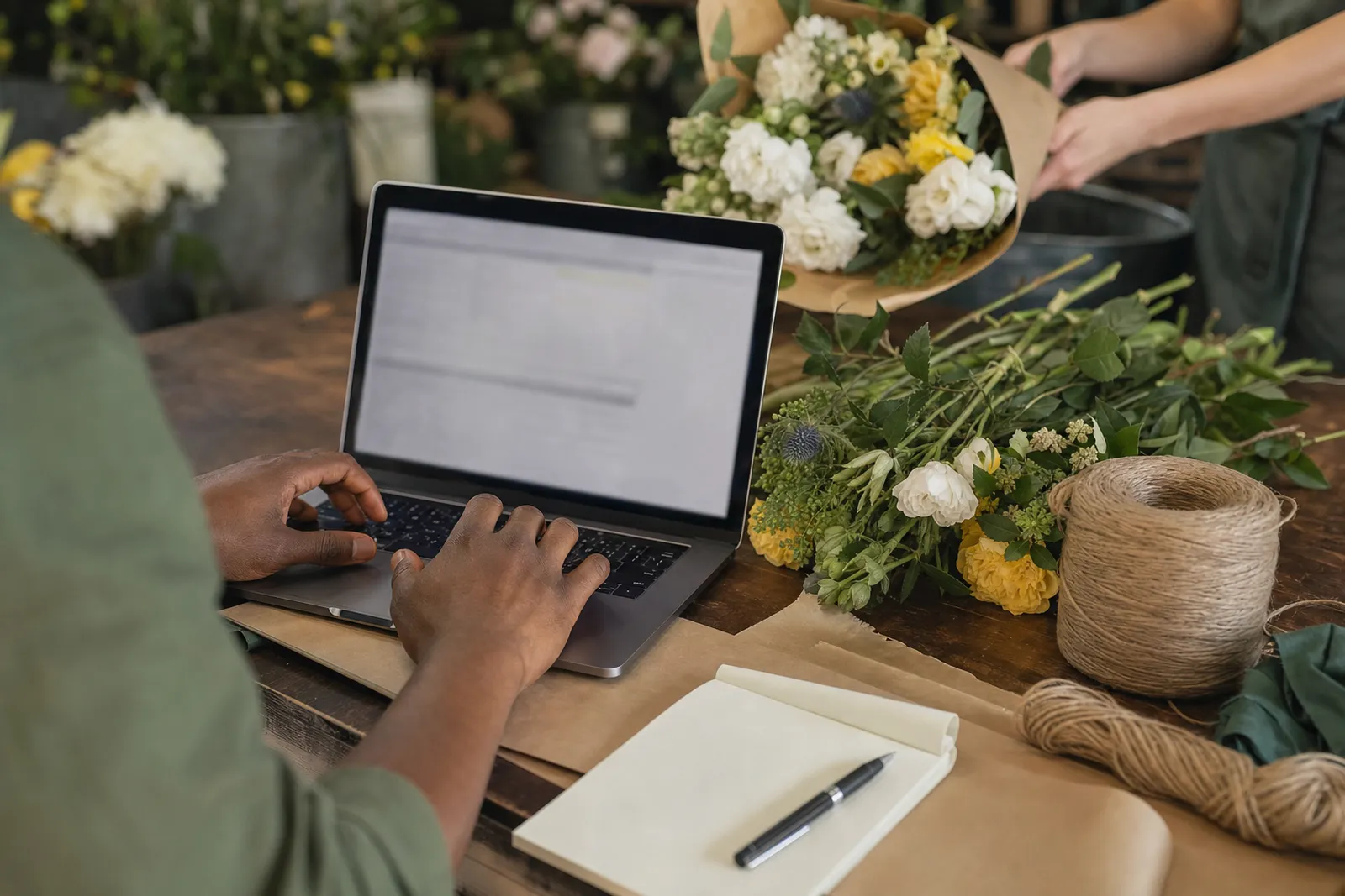 Hands of a florist typing on a laptop beside bundles of cut flowers while a colleague passes a bouquet