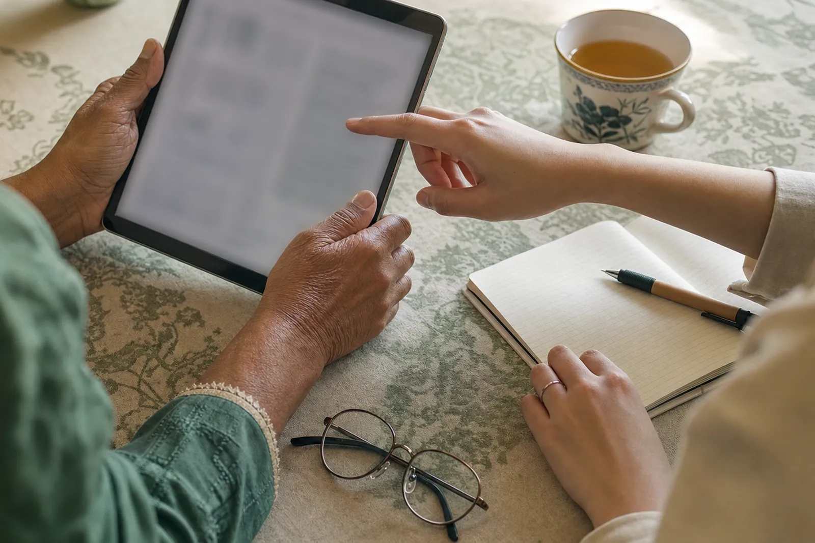 Older hands holding a tablet at a kitchen table while a younger person points at the screen, with reading glasses, tea and a notebook nearby